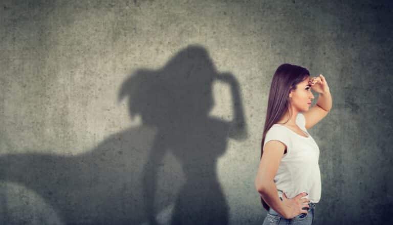 Frau steht vor einer Wand, deren Schatten ein wehendes Helden-Cape zeigt – Metapher für das Helfersyndrom in der Beziehung und emotionale Abhängigkeit.